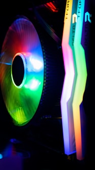 A colorful computer cooling fan alongside RGB illuminated RAM sticks inside a computer case. The fan showcases a vibrant spectrum of colors including green, blue, purple, and yellow, while the RAM sticks exhibit a glowing gradient of red, green, yellow, and blue hues.