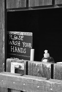 A monochrome image shows a sign with the words 'Please wash your hands,' partially obscured by a wooden window or stall door. A bottle of liquid soap with a pump is also visible through the opening.