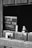 A monochrome image shows a sign with the words 'Please wash your hands,' partially obscured by a wooden window or stall door. A bottle of liquid soap with a pump is also visible through the opening.