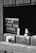 A monochrome image shows a sign with the words 'Please wash your hands,' partially obscured by a wooden window or stall door. A bottle of liquid soap with a pump is also visible through the opening.