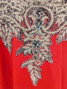 A detailed embroidery pattern featuring intricate floral and leaf motifs adorned with sparkling beads and sequins. The design is set against a vibrant red fabric background, showcasing a blend of gold and black hues in the embellishments.