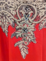 A detailed embroidery pattern featuring intricate floral and leaf motifs adorned with sparkling beads and sequins. The design is set against a vibrant red fabric background, showcasing a blend of gold and black hues in the embellishments.
