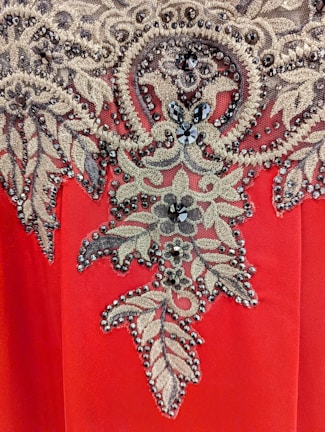 A detailed embroidery pattern featuring intricate floral and leaf motifs adorned with sparkling beads and sequins. The design is set against a vibrant red fabric background, showcasing a blend of gold and black hues in the embellishments.