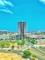 A partially constructed high-rise building stands in an urban area, surrounded by roads and green spaces. Several cars are traveling on the roads nearby, and trees provide a natural touch to the otherwise industrial setting. The sky is clear and blue, adding to the open and airy feel of the location.