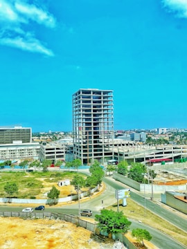 A partially constructed high-rise building stands in an urban area, surrounded by roads and green spaces. Several cars are traveling on the roads nearby, and trees provide a natural touch to the otherwise industrial setting. The sky is clear and blue, adding to the open and airy feel of the location.