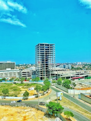 A partially constructed high-rise building stands in an urban area, surrounded by roads and green spaces. Several cars are traveling on the roads nearby, and trees provide a natural touch to the otherwise industrial setting. The sky is clear and blue, adding to the open and airy feel of the location.
