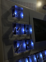 A wall-mounted display features illuminated boxes, each containing chemical element samples such as Hydrogen, Lithium, Beryllium, and more. The boxes emit a soft blue light, creating a modern and scientific ambiance.