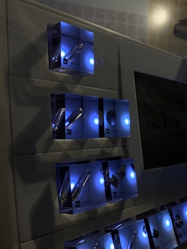 A wall-mounted display features illuminated boxes, each containing chemical element samples such as Hydrogen, Lithium, Beryllium, and more. The boxes emit a soft blue light, creating a modern and scientific ambiance.