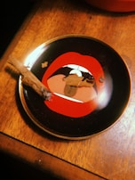 A wooden surface with a round black ashtray featuring a red open mouth design. The ashtray contains tobacco remnants, ash, and a partially smoked cigarillo resting on its edge.