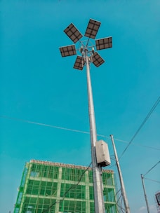 A tall street light with multiple solar panels is prominently displayed against a clear blue sky. In the background, there is a partially constructed building covered with green safety netting. Several electrical wires are also visible.