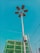 A tall street light with multiple solar panels is prominently displayed against a clear blue sky. In the background, there is a partially constructed building covered with green safety netting. Several electrical wires are also visible.
