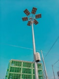 A tall street light with multiple solar panels is prominently displayed against a clear blue sky. In the background, there is a partially constructed building covered with green safety netting. Several electrical wires are also visible.