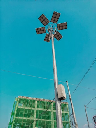 A tall street light with multiple solar panels is prominently displayed against a clear blue sky. In the background, there is a partially constructed building covered with green safety netting. Several electrical wires are also visible.