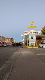 A vintage motor hotel sign with Route 66 insignia stands on a street lined with parked cars and historic-style buildings, set during the early evening. The street is illuminated by lampposts and the sky has a faint blue hue.