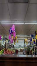 A vibrant supermarket aisle decorated with colorful piñatas in pink, purple, and yellow hanging from the ceiling. Fresh produce like apples, pineapples, and other fruits are neatly arranged on shelves. The area is well-lit with fluorescent lighting, and there are various packaged goods on the shelves.