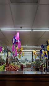 A vibrant supermarket aisle decorated with colorful piñatas in pink, purple, and yellow hanging from the ceiling. Fresh produce like apples, pineapples, and other fruits are neatly arranged on shelves. The area is well-lit with fluorescent lighting, and there are various packaged goods on the shelves.