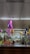 A vibrant supermarket aisle decorated with colorful piñatas in pink, purple, and yellow hanging from the ceiling. Fresh produce like apples, pineapples, and other fruits are neatly arranged on shelves. The area is well-lit with fluorescent lighting, and there are various packaged goods on the shelves.