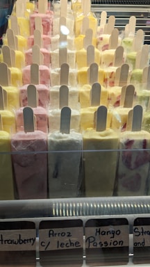 A display case filled with various colorful ice cream popsicles arranged in rows. Each popsicle is individually wrapped and has a wooden stick. Flavors include strawberry, arroz con leche, mango passion, and others with vibrant colors ranging from yellow to pink.