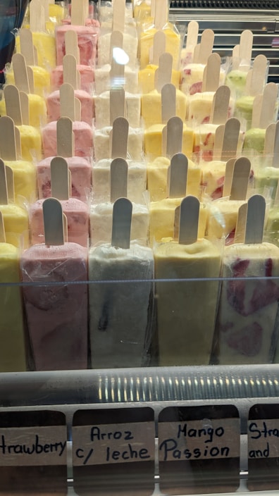 A display case filled with various colorful ice cream popsicles arranged in rows. Each popsicle is individually wrapped and has a wooden stick. Flavors include strawberry, arroz con leche, mango passion, and others with vibrant colors ranging from yellow to pink.