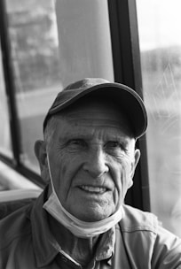 An elderly man with a cap is sitting by a window. He has a gentle smile and is wearing a face mask pulled under his chin. The setting appears to be indoors with natural light coming from the window.