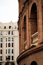 An architectural scene featuring a red brick building with arched windows and decorative stone elements in the foreground. In the background, a large white traditional building with symmetrical window placements can be seen. A vintage-style streetlamp adds character to the urban setting.