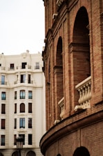 An architectural scene featuring a red brick building with arched windows and decorative stone elements in the foreground. In the background, a large white traditional building with symmetrical window placements can be seen. A vintage-style streetlamp adds character to the urban setting.