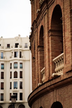 An architectural scene featuring a red brick building with arched windows and decorative stone elements in the foreground. In the background, a large white traditional building with symmetrical window placements can be seen. A vintage-style streetlamp adds character to the urban setting.