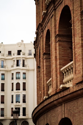 An architectural scene featuring a red brick building with arched windows and decorative stone elements in the foreground. In the background, a large white traditional building with symmetrical window placements can be seen. A vintage-style streetlamp adds character to the urban setting.