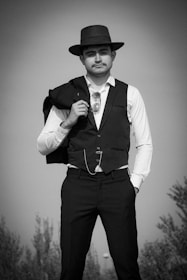 A man is dressed in a formal attire, wearing a vest and a fedora hat. He holds a jacket over his shoulder, and a pair of sunglasses is tucked into his vest pocket. He exudes a classic, vintage style with a confident posture.