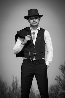 A man is dressed in a formal attire, wearing a vest and a fedora hat. He holds a jacket over his shoulder, and a pair of sunglasses is tucked into his vest pocket. He exudes a classic, vintage style with a confident posture.