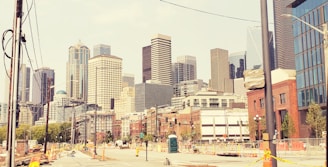A cityscape with a variety of skyscrapers displaying modern and classical architecture styles. The street in the foreground is under construction with visible barriers, cones, and a portable toilet. The scene includes trees and street lamps along the sidewalk.