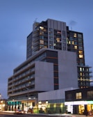 A modern, multi-story residential or commercial building with large windows and balconies is illuminated in the evening. The structure features different geometric sections with contrasting colors, primarily gray, black, and orange accents. Below, street-level shops are visible with bright signage, and there is sparse foot traffic and few vehicles on the street.