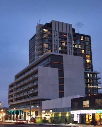 A modern, multi-story residential or commercial building with large windows and balconies is illuminated in the evening. The structure features different geometric sections with contrasting colors, primarily gray, black, and orange accents. Below, street-level shops are visible with bright signage, and there is sparse foot traffic and few vehicles on the street.