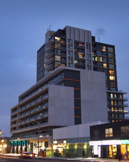 A modern, multi-story residential or commercial building with large windows and balconies is illuminated in the evening. The structure features different geometric sections with contrasting colors, primarily gray, black, and orange accents. Below, street-level shops are visible with bright signage, and there is sparse foot traffic and few vehicles on the street.