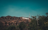 The image features the iconic Hollywood sign situated on a hilly landscape with a communication tower in the background. Trees frame the scene, with a clear sky overhead.