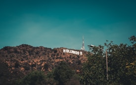 The image features the iconic Hollywood sign situated on a hilly landscape with a communication tower in the background. Trees frame the scene, with a clear sky overhead.