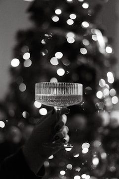 A hand holds a delicate crystal glass filled with a transparent liquid against a backdrop of blurred, glowing lights. The elegant features of the glass stand out in the soft monochrome palette.