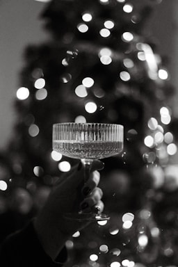 A hand holds a delicate crystal glass filled with a transparent liquid against a backdrop of blurred, glowing lights. The elegant features of the glass stand out in the soft monochrome palette.