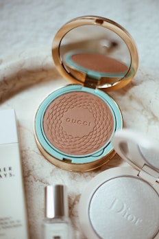 A luxurious arrangement of cosmetic products featuring a Gucci bronzer in an elegantly designed mirrored compact, surrounded by additional beauty items including a Dior compact and a small glass bottle. The setting has a sophisticated and stylish aesthetic with a marble surface.