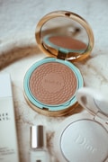 A luxurious arrangement of cosmetic products featuring a Gucci bronzer in an elegantly designed mirrored compact, surrounded by additional beauty items including a Dior compact and a small glass bottle. The setting has a sophisticated and stylish aesthetic with a marble surface.