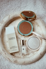 A collection of luxury beauty products is arranged on a textured, light cream surface. There is a bronzing powder with a turquoise and gold compact labeled 'Gucci', a bottle of 'Summer Fridays' mineral milk sunscreen, a small bottle of Jo Malone perfume, and a Dior highlighter in a white, round case.