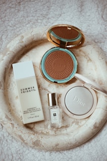 A collection of luxury beauty products is arranged on a textured, light cream surface. There is a bronzing powder with a turquoise and gold compact labeled 'Gucci', a bottle of 'Summer Fridays' mineral milk sunscreen, a small bottle of Jo Malone perfume, and a Dior highlighter in a white, round case.