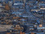 An overhead view of a rural village featuring numerous wooden houses clustered alongside a winding road. Around the houses are haystacks and bare trees, giving an impression of a late autumn or early spring scene. The colors are muted, with a dominance of browns and grays.
