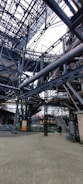 An intricate network of metal beams and structures form a complex industrial setting. The steel framework dominates the scene with staircases and platforms visible. A person stands on the ground, giving a sense of scale to the massive construction.