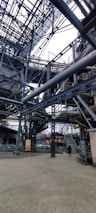 An intricate network of metal beams and structures form a complex industrial setting. The steel framework dominates the scene with staircases and platforms visible. A person stands on the ground, giving a sense of scale to the massive construction.
