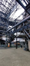 An intricate network of metal beams and structures form a complex industrial setting. The steel framework dominates the scene with staircases and platforms visible. A person stands on the ground, giving a sense of scale to the massive construction.