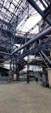 An intricate network of metal beams and structures form a complex industrial setting. The steel framework dominates the scene with staircases and platforms visible. A person stands on the ground, giving a sense of scale to the massive construction.