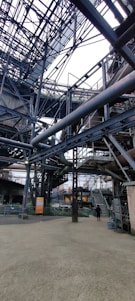 An intricate network of metal beams and structures form a complex industrial setting. The steel framework dominates the scene with staircases and platforms visible. A person stands on the ground, giving a sense of scale to the massive construction.
