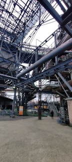 An intricate network of metal beams and structures form a complex industrial setting. The steel framework dominates the scene with staircases and platforms visible. A person stands on the ground, giving a sense of scale to the massive construction.