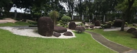 A peaceful garden scene with large, smooth boulders scattered across a well-manicured lawn. A narrow path winds through the area, flanked by neatly trimmed shrubs and lush greenery. Tall trees at the back provide a sense of enclosure, while sunlight filters through the leaves, casting dappled shadows on the ground.
